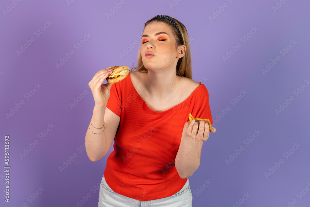 stained smeared in mayonnaise pretty girl eats fast food, a cheap burger, holds cheeseburger. fast food and unhealthy diet. isolated on purple background. stained smeared in mayonnaise pretty girl eats fast food, a cheap burger, holds cheeseburger. fast food and unhealthy diet. isolated on purple background.