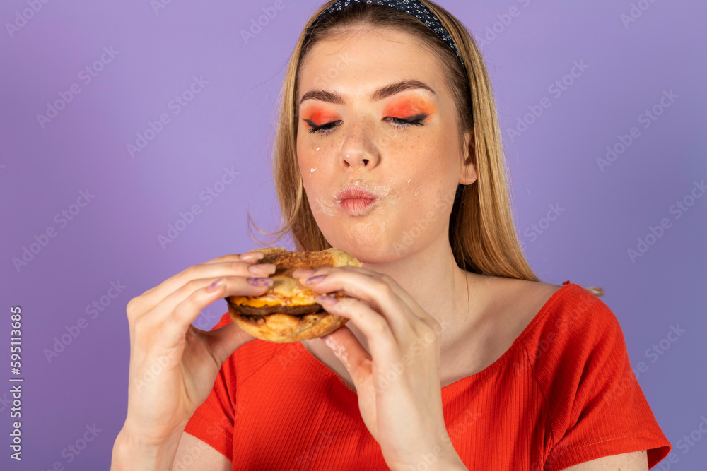 stained smeared in mayonnaise pretty girl eats fast food, a cheap burger, holds cheeseburger. fast food and unhealthy diet. isolated on purple background. stained smeared in mayonnaise pretty girl eats fast food, a cheap burger, holds cheeseburger. fast food and unhealthy diet. isolated on purple background.