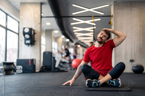 Young male athlete doing stretching exercise in gym while sitting with crossed legs on exercise mat. Sportsman in red t-shirt stretching neck muscles during warm up session, sitting on floor in gym.