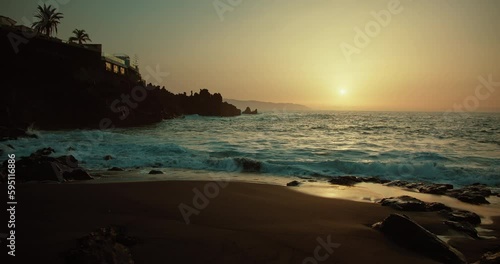 Ocean waves roll in slow-motion on black sandy smooth beach at sunset. Sea water splashing at dusk evening cloudy sky. photo