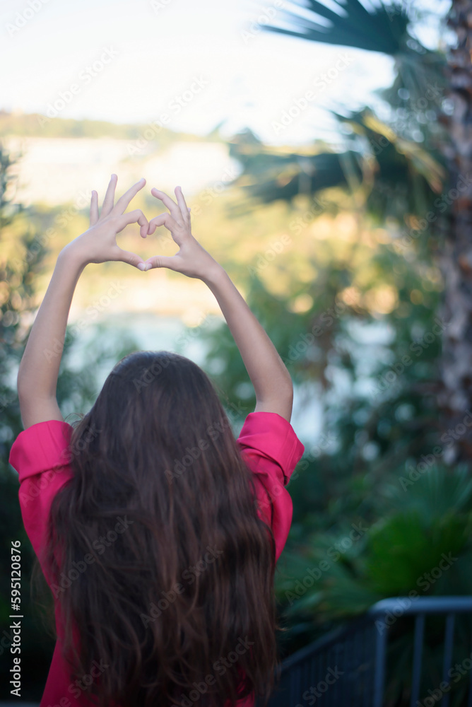 Obraz premium A view of the back of a heart shaped hands of young girl with long hair, pink shirt. Heart made by kids hand at sunset. Tropical view, palms around