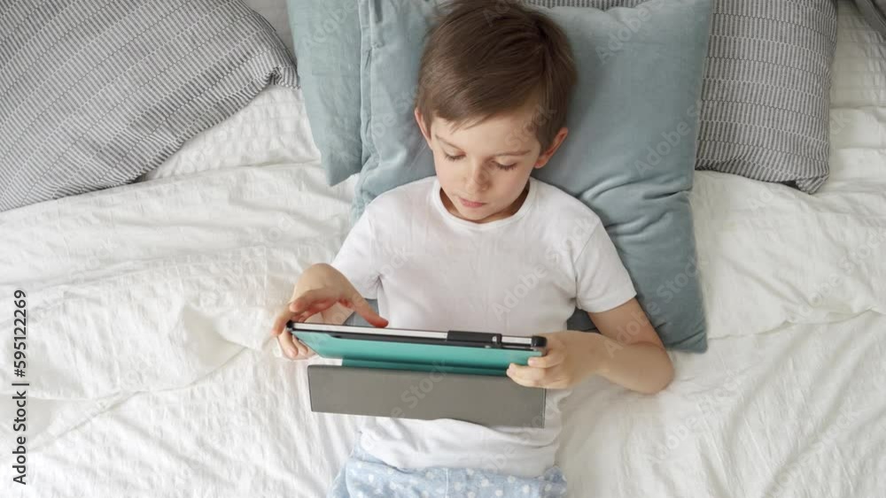 A boy using a tablet computer while lying in bed during the day. His gaze is fixed on the screen. Child using gadget, education and development