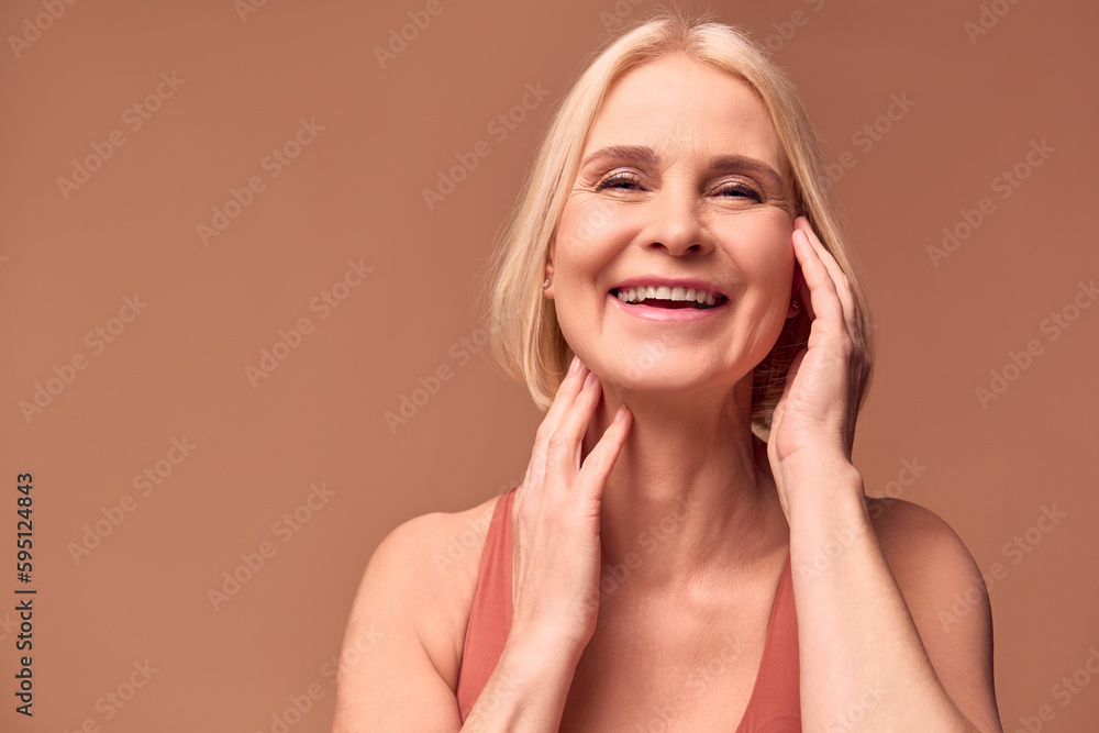A cheerful elderly woman touches the skin of her face with her hands and laughs. The concept of skin care, female beauty.Beige background. A cheerful elderly woman touches the skin of her face with her hands and laughs. The concept of skin care, female beauty.Beige background.