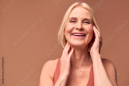 A cheerful elderly woman touches the skin of her face with her hands and laughs. The concept of skin care, female beauty.Beige background.
