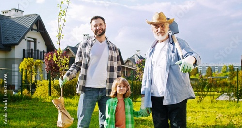 Portrait shot of cute small boy with happy father and grandfather smiling to camera in garden. Male genarations of family planting trees at summerhouse. Outside. photo