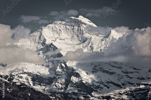 In this stunning image, a majestic mountain peak rises above the clouds on a beautiful sunny day in Interlaken, Switzerland, creating a captivating and peaceful scene.