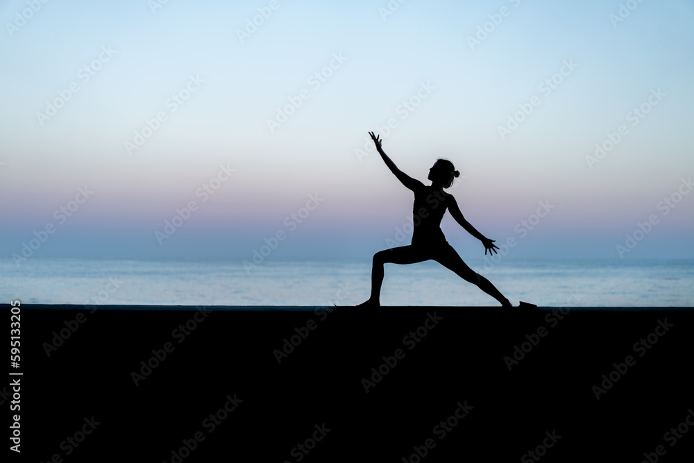Silhouette de femme faisant du yoga devant la mer au coucher de soleil,  posture Virabhadrasana (Warrior - Guerrier)