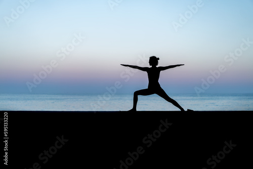 Silhouette de femme faisant du yoga devant la mer au coucher de soleil, posture Virabhadrasana II (Warrior II - Guerrier II)