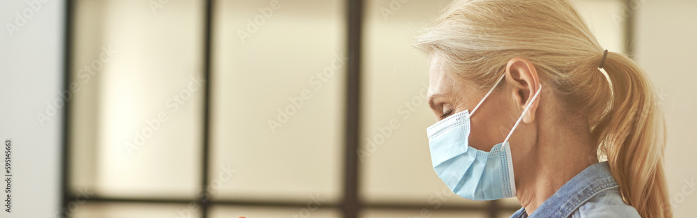 Side view of mature blonde woman in protective face mask rubbing, cleaning her hands using antibacterial hand sanitizer, standing indoors Side view of mature blonde woman in protective face mask rubbing, cleaning her hands using antibacterial hand sanitizer, standing indoors