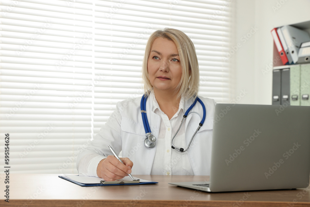Doctor with laptop and clipboard writing at wooden table in clinic Doctor with laptop and clipboard writing at wooden table in clinic