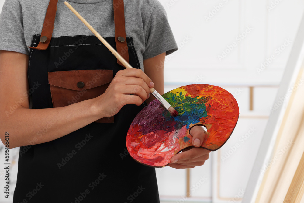 Fototapeta premium Woman mixing paints on palette with brush near easel in studio, closeup