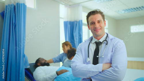Portrait of male doctor looking to camera and smile feel happy. Background patient in hospital room. Positive general practitioner close up face portrait.