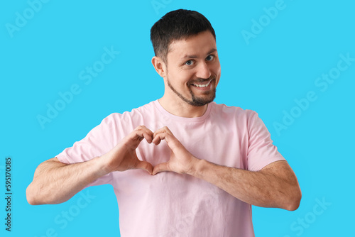 Handsome man in t-shirt making heart shape with hands on blue background