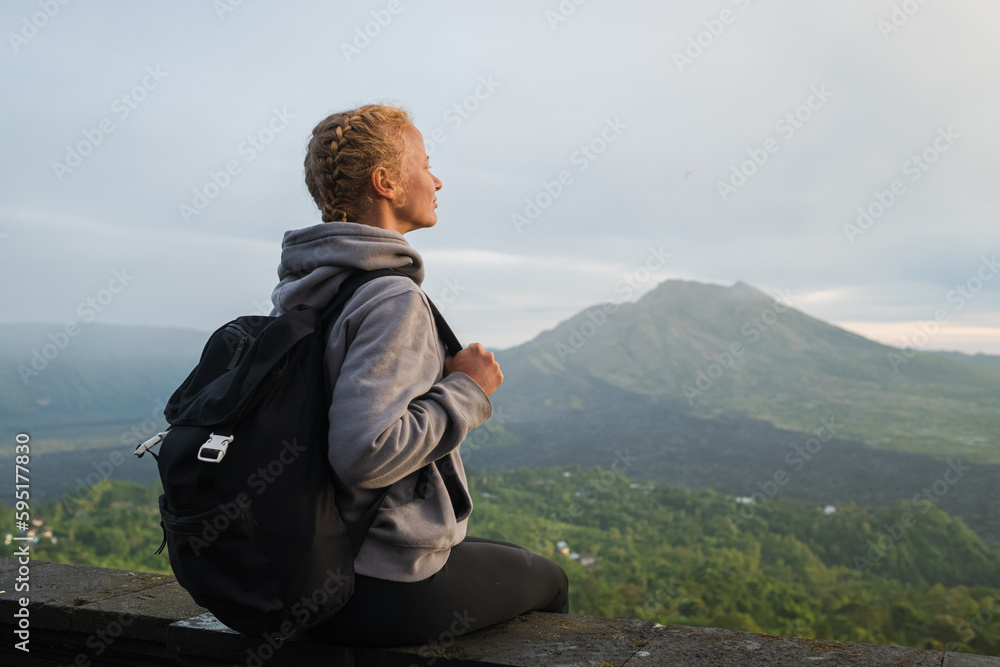 A girl traveler with a backpack walks in the mountains at dawn and looks at the Batur volcano on the island of Bali, Indonesia. A girl traveler with a backpack walks in the mountains at dawn and looks at the Batur volcano on the island of Bali, Indonesia.