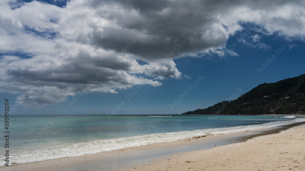 The waves of the turquoise ocean are foaming on the beach. Wet sand glistens. A green hill against a background of blue sky and clouds. Seychelles. Mahe. Beau Vallon