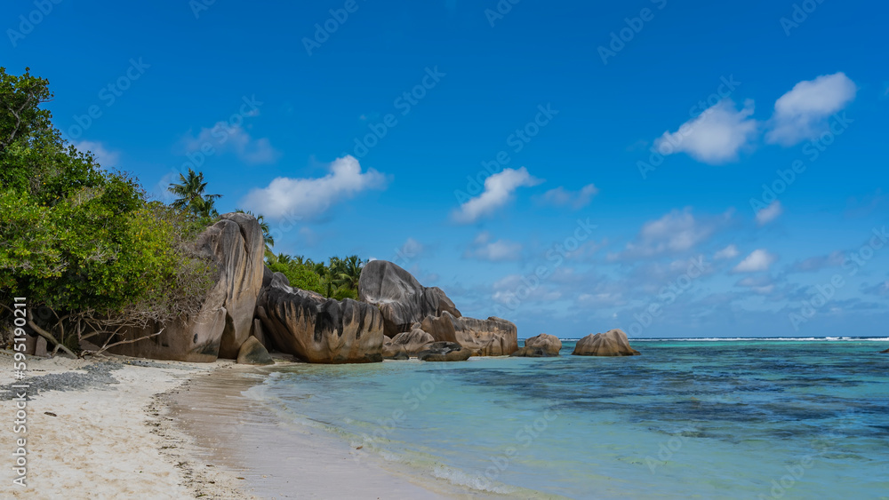 Picturesque granite boulders and rocks with steep smoothed slopes on a tropical beach. Turquoise ocean waves on the sand. Lush tropical vegetation. Blue sky, clouds. Seychelles. La Digue. 