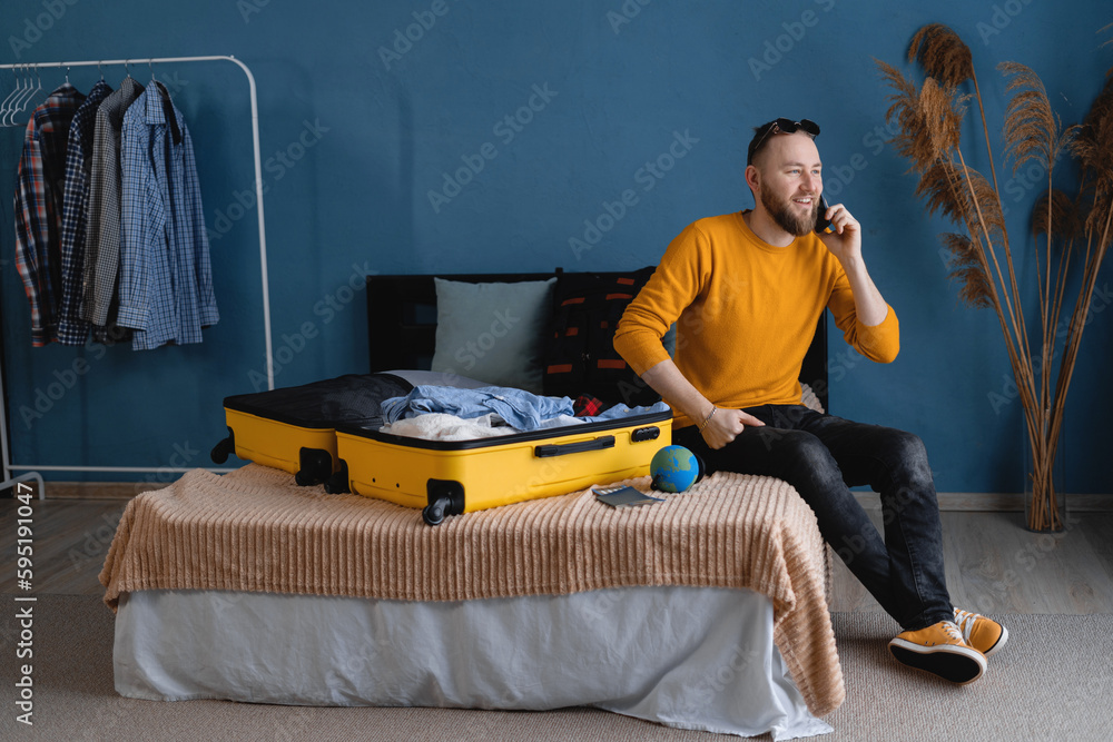 Young travel man sitting on bed near open suitcase with clothes, speaking on smartphone, booking travel tour at home, hotel reservation, buying tickets on phone.