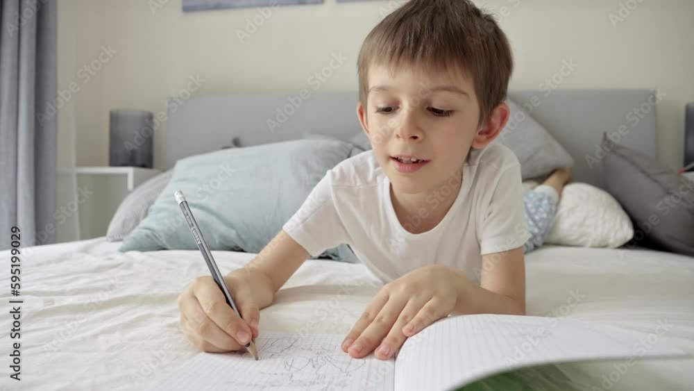 Young boy in pajamas writing in a notebook while lying in bed, doing his homework. Education and child development in a remote learning environment Young boy in pajamas writing in a notebook while lying in bed, doing his homework. Education and child development in a remote learning environment