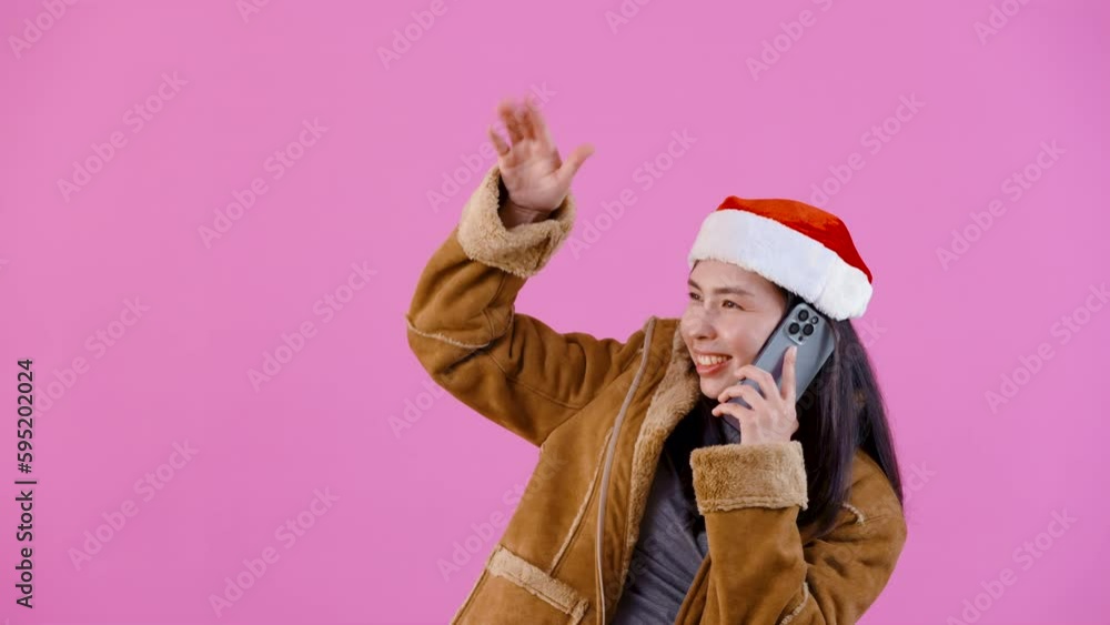 4K, Beautiful Asian girl in long-sleeved shirt wearing Christmas hat, Talking on phone, looking for another person, making gestures waving for others to see, Isolated indoor studio on pink background. 4K, Beautiful Asian girl in long-sleeved shirt wearing Christmas hat, Talking on phone, looking for another person, making gestures waving for others to see, Isolated indoor studio on pink background.