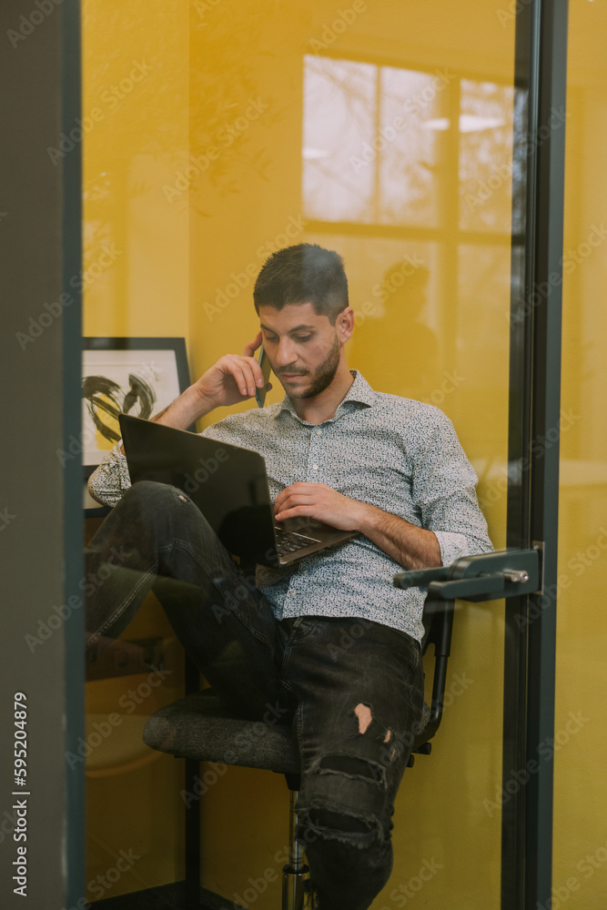 Good looking male business person having phone call in soundproof phone booth at the office while working on his lap top Good looking male business person having phone call in soundproof phone booth at the office while working on his lap top