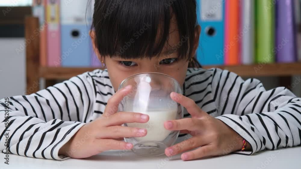 Cute Asian girl drinking a glass of milk at home in living room. Little girl drinking milk in the morning before going to school. Healthy food in childhood. Cute Asian girl drinking a glass of milk at home in living room. Little girl drinking milk in the morning before going to school. Healthy food in childhood.
