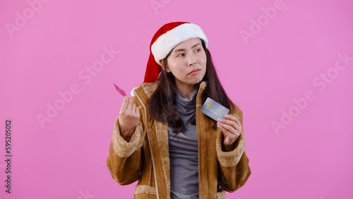 4K, Beautiful Asian girl in long-sleeved shirt wearing Christmas hat, She is holding two credit cards, red and gray, thinking about going shopping, Isolated indoor studio on pink background. photo