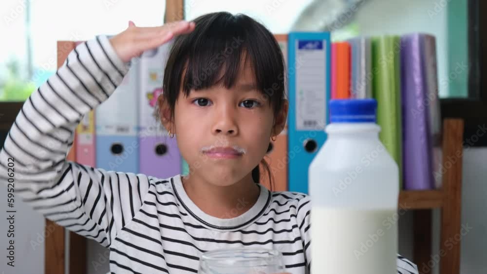 Cute Asian girl drinking a glass of milk at home in living room. Little girl drinking milk in the morning before going to school. Healthy food in childhood. Cute Asian girl drinking a glass of milk at home in living room. Little girl drinking milk in the morning before going to school. Healthy food in childhood.