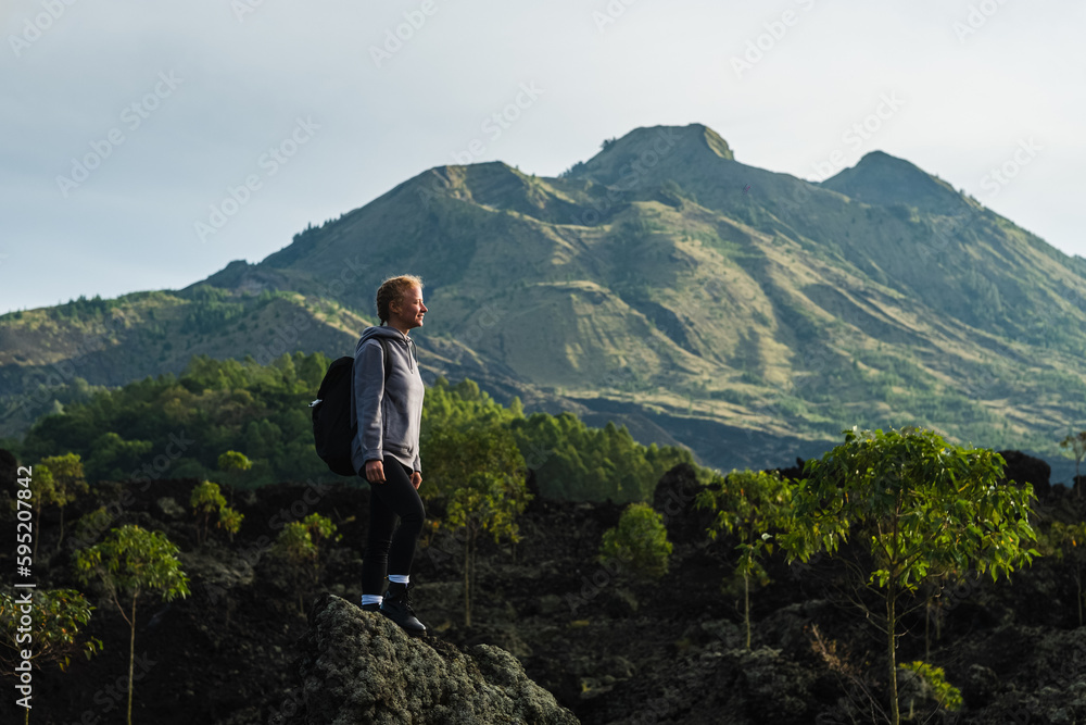 Fototapeta premium A young girl traveler with a backpack walks in the mountains against the backdrop of the Batur volcano on the island of Bali in Indonesia.
