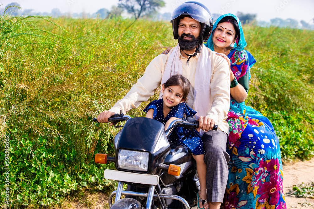 Happy young rural Indian family wearing safety helmet riding on motorcycle in village agricultural field. Mode of transportation, Copy space