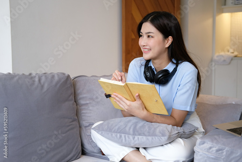 Bright and cute Asian woman sitting on sofa with laptop to work and happily taking notes at home.