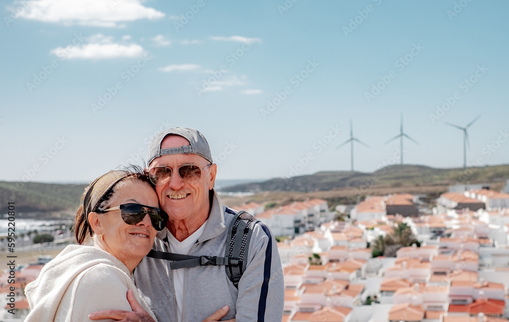 Loving old senior family couple bonding embracing enjoying sunny day in outdoors. Windmill on background. Elderly people in vacation or retirement at sea
