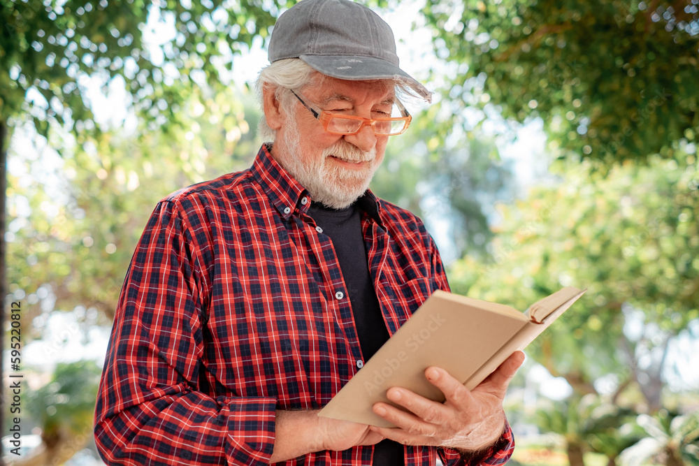 Portrait of senior bearded man with hat reading a book sitting in the public park. Elderly relaxed pensioner with eyeglasses enjoying retirement in outdoor Portrait of senior bearded man with hat reading a book sitting in the public park. Elderly relaxed pensioner with eyeglasses enjoying retirement in outdoor