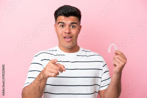 Young caucasian man holding invisaling isolated on pink background surprised and pointing front photo