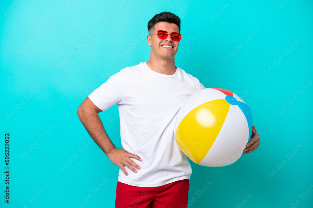 Young caucasian man holding a beach ball isolated on blue background posing with arms at hip and smiling