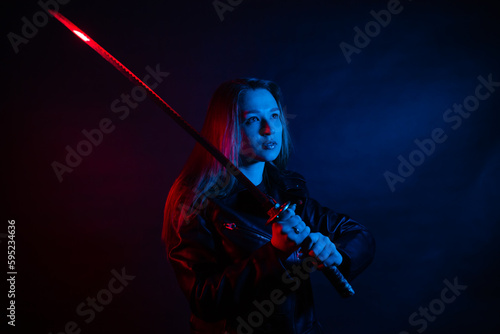 A young beautiful woman with long hair in a black leather jacket holds a katana at night in the dark. Robber in the city in neon blue and red light with melee weapons.