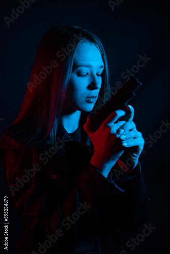 A young beautiful woman with long hair in a black leather jacket is holding a gun in the dark at night. Robber in the city in neon blue and red light.