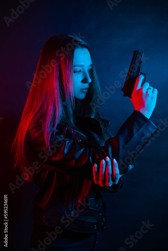 A young beautiful woman with long hair in a black leather jacket is holding a gun in the dark at night. Robber in the city in neon blue and red light.