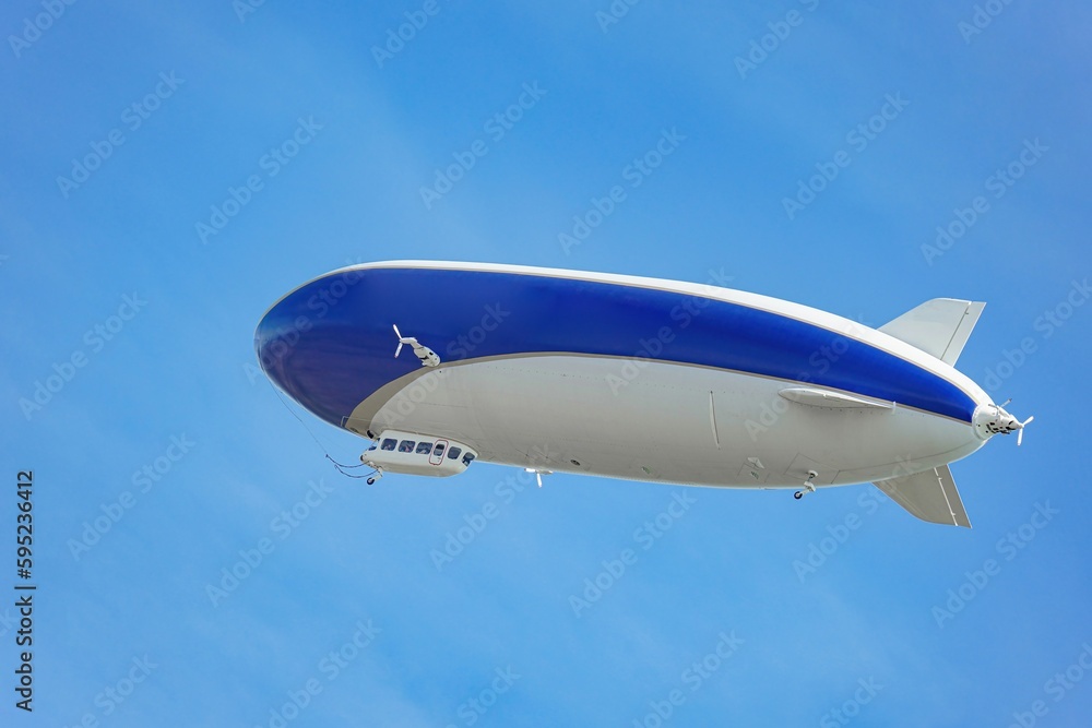 A blue and white airship with passengers on board flies against the blue sky. A blue and white airship with passengers on board flies against the blue sky.