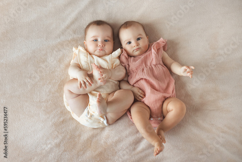 Top view of two cute baby twin girls 6 month old lying together on comfortable and soft bed with beige cotton blanket at home

