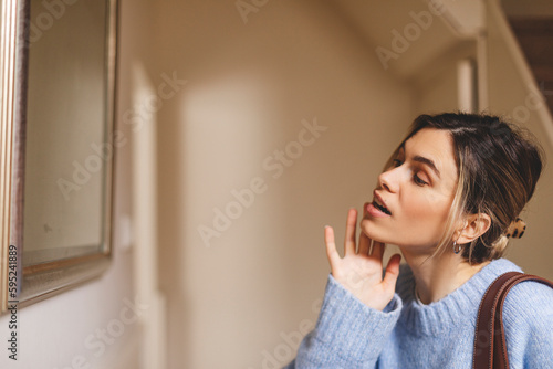 Side view of pretty young woman touch her face before go away from home. Girl prepare before leaving home. Woman wear blue knitted sweater.
