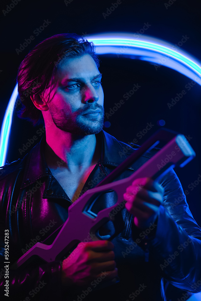 Handsome man in cyberpunk style with a machine gun in his hands in the dark. A guy in a leather jacket in the scenery of the future in neon color.