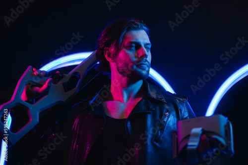 Handsome man in cyberpunk style with a machine gun in his hands in the dark. A guy in a leather jacket in the scenery of the future in neon color.