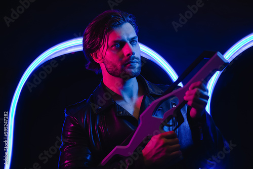 Handsome man in cyberpunk style with a machine gun in his hands in the dark. A guy in a leather jacket in the scenery of the future in neon color.