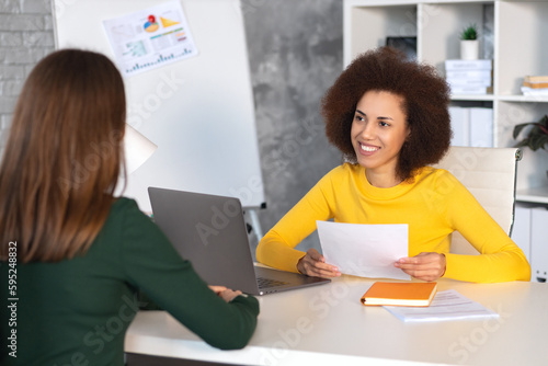 Friendly interview between African American businesswoman hr director and caucasian job candidate. Recruitment manager holding paper cv and talking, hiring for job female sitting in office