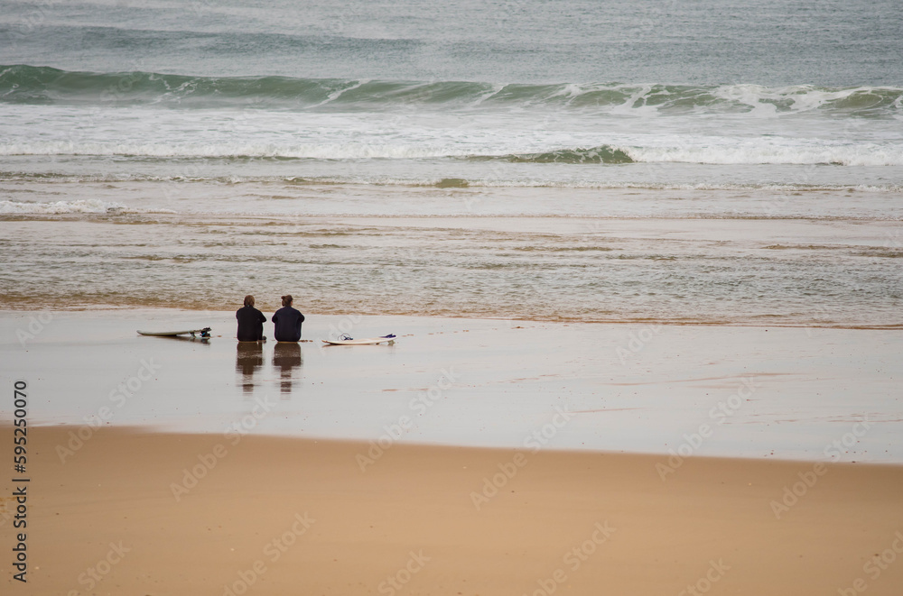 Couple of surfers sitting on the sand facing the Atlantic Ocean Couple of surfers sitting on the sand facing the Atlantic Ocean