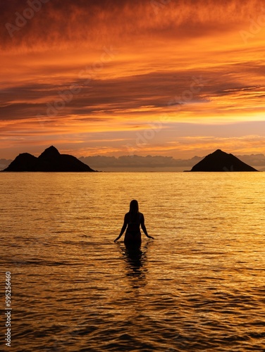 Woman walking in the ocean toward the horizon as the sun sets and two islands in the background