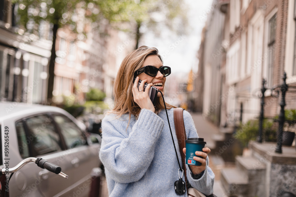 Fototapeta premium Beautiful young woman wearing blue sweater, walking outdoors, talking on mobile phone. Beautiful stylish woman talking on phone, calling on mobile phone walking outdoor, hold coffee cup take away.