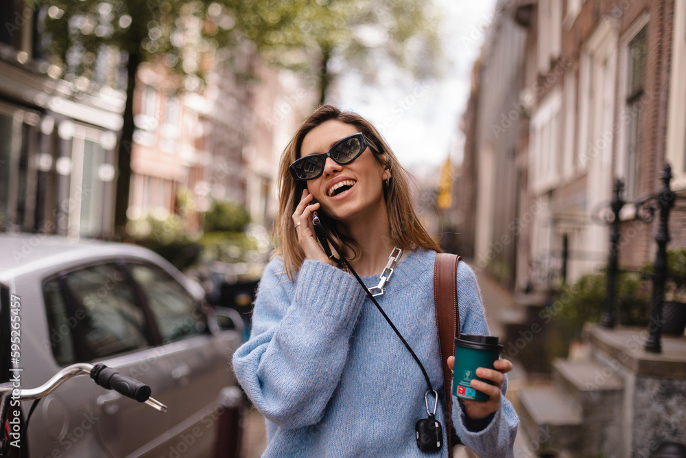 Fototapeta premium Beautiful young woman wearing blue sweater, walking outdoors, talking on mobile phone. Beautiful stylish woman talking on phone, calling on mobile phone walking outdoor, hold coffee cup take away.