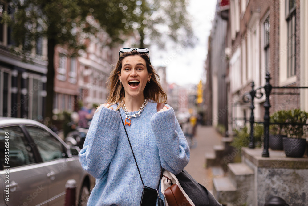 Delighted blonde girl making winner gesture and using mobile phone walking on the street. Portrait of surprised hipster girl outdoors. Girl wear blue sweater, black sunglasses, bag and look happy.