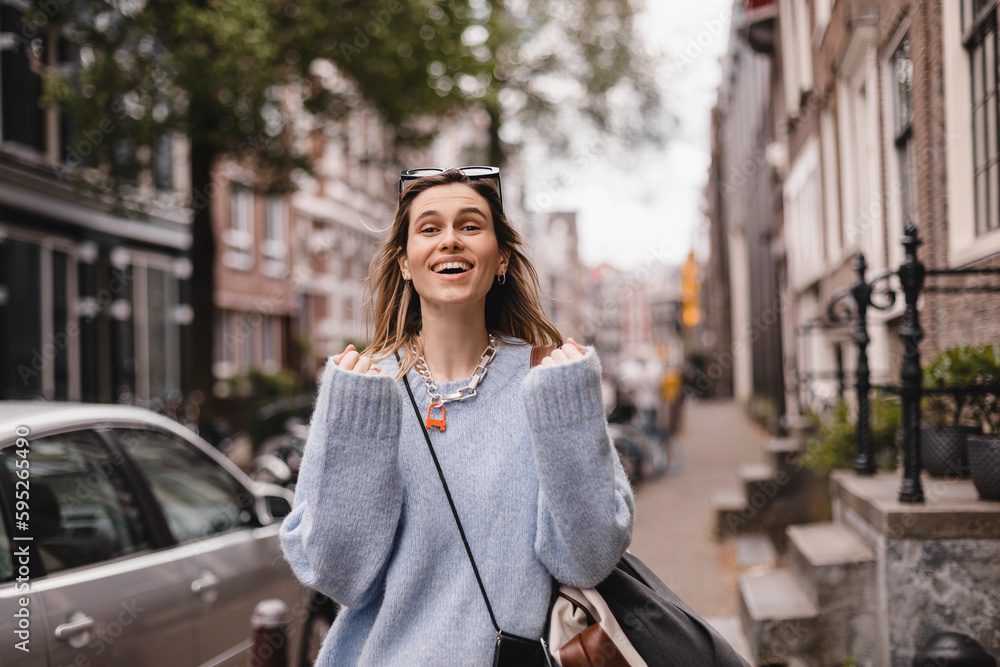 Delighted blonde girl making winner gesture and using mobile phone walking on the street. Portrait of surprised hipster girl outdoors. Girl wear blue sweater, black sunglasses, bag and look happy.